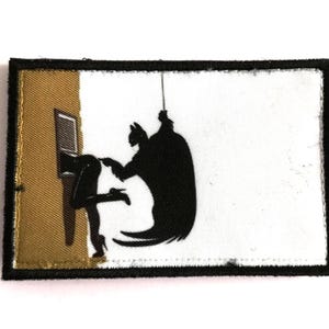 Pode incluir: Um patch retangular com uma borda preta. O patch apresenta uma silhueta preta de uma figura semelhante ao Batman interagindo com uma mulher. O fundo é branco e dourado. O patch é provavelmente para passar a ferro ou costurar.