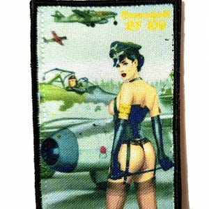 Könnte beinhalten: Gestickter Aufnäher mit einem Vintage-Pin-up-Girl-Design in militärischem Ambiente. Das Design zeigt eine Frau in einem blauen Korsett und schwarzen Handschuhen, mit Flugzeugen im Hintergrund und dem Text "Dornier 87 KG".
