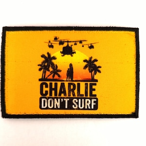Peut inclure: Écusson rectangulaire avec un fond jaune et une bordure noire. L'écusson présente une silhouette de palmiers, d'hélicoptères et le texte "CHARLIE DON'T SURF" en lettres blanches.