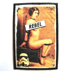 Könnte beinhalten: Gestickter Aufnäher mit einem Pin-up-Stil-Bild einer Frau und dem Wort "REBEL" auf einem Schild. Ein Stormtrooper-Helm ist ebenfalls vorhanden. Der Aufnäher hat einen schwarzen Rand und eine Vintage-Ästhetik.