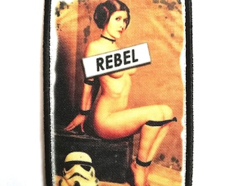 May include: Embroidered patch featuring a pin-up style image of a woman with the word "REBEL" on a sign. The patch includes a Stormtrooper helmet and a black border. The image has a vintage aesthetic.