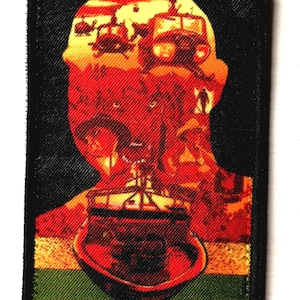 Peut inclure: Un écusson rectangulaire avec une bordure noire. L'écusson présente une silhouette de tête remplie d'une image rouge, orange et jaune d'hélicoptères et d'un bateau. Le bas de l'écusson est vert et jaune.
