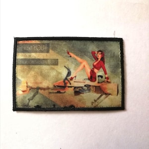 Könnte beinhalten: Rechteckiger Aufnäher mit einer Pin-up-Girl-Illustration. Die Frau, in roter Kleidung, liegt auf einer Maschine. Der Text oben lautet "SHE WANT YOU!" und darunter "HEROES BE SCOUTED OVER". Der Aufnäher hat einen schwarzen Rand.
