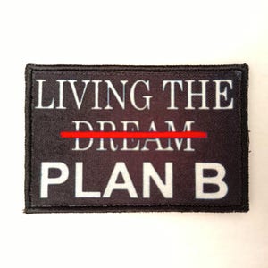 Peut inclure: Écusson rectangulaire noir avec le texte blanc « LIVING THE DREAM » avec une ligne rouge traversant le mot « DREAM » et le texte « PLAN B » en dessous. L'écusson a une bordure noire.