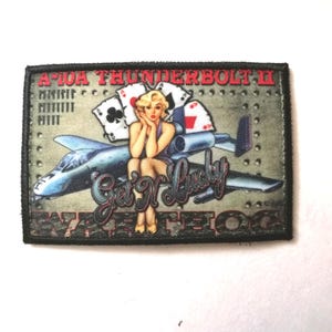 May include: Rectangular patch featuring a pin-up girl, playing cards, and an A-10A Thunderbolt II aircraft. The patch has the text "A-10A THUNDERBOLT II", "Get N Lucky", and "Warthog". The background is olive green.