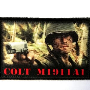 May include: Rectangular patch featuring a soldier aiming a handgun. The image has a blurred background of trees. The text "COLT M1911A1" is printed in red at the bottom of the patch. The patch has a black border.