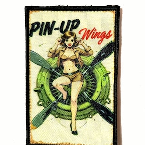 Peut inclure: Un écusson rectangulaire avec une illustration de pin-up. L'image présente une femme en tenue marron avec les mots "PIN-UP Wings" en noir et rouge. Une hélice et un moteur verts sont en arrière-plan.