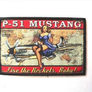 May include: Rectangular patch with a black border featuring a vintage illustration. The design includes a P-51 Mustang airplane, a woman in a blue dress, and the text "P-51 Mustang" and "Fire the Rockets, Baby!" against a map background.