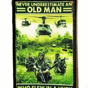 May include: Embroidered patch with the text "NEVER UNDERESTIMATE AN OLD MAN WHO FLEW IN A HUEY." The patch features a green background with helicopters and soldiers. The patch has a black border.