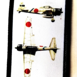 Puede incluir: Un parche rectangular con borde negro presenta dos ilustraciones de un avión de combate japonés Zero. Los aviones son de color crema con insignias circulares rojas y detalles negros. Un avión se ve desde arriba, el otro de lado.