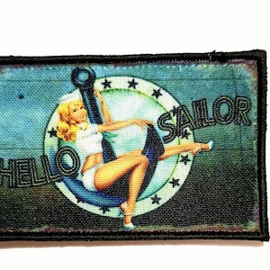 May include: Rectangular patch with a vintage pin-up girl design. The image features a blonde woman in a sailor outfit, seated on an anchor. The words "HELLO SAILOR" are printed on the patch. The patch has a black border.