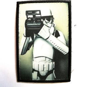 Peut inclure: Écusson rectangulaire avec une bordure noire représentant un Stormtrooper de Star Wars tenant un appareil photo vintage. Le fond est vert et blanc délavé. Le Stormtrooper est en armure blanche.