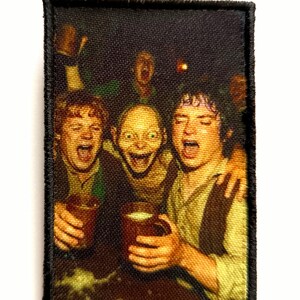 Può includere: Una toppa rettangolare con bordo nero raffigura una scena di un film. Tre personaggi sono mostrati con la bocca aperta, che tengono boccali di birra. L'immagine ha uno sfondo scuro e un aspetto strutturato.