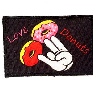 Op de afbeelding: Zwart rechthoekig embleem met de woorden "Love Donuts" in roze en rood. Het embleem toont twee donuts, een roze en een rode, en een witte cartoonhand die een vredesteken maakt.
