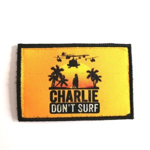 Può includere: Toppa rettangolare con bordo nero e sfondo giallo e arancione. La toppa presenta una silhouette di elicottero, palme e una persona con la scritta "CHARLIE DON'T SURF" in nero.