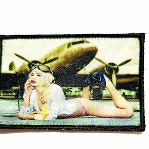 Könnte beinhalten: Gestickter Aufnäher mit einem Vintage-Bild einer Frau in Fliegerkleidung, die vor einem Propellerflugzeug liegt. Der Aufnäher hat einen schwarzen Rand und eine Retro-Ästhetik mit gedämpften Farben.