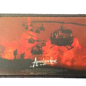Può includere: Toppa rettangolare con una scena del film "Apocalypse Now". L'immagine raffigura elicotteri contro un cielo rosso fuoco, con il titolo del film in scrittura bianca. La toppa ha un bordo nero.