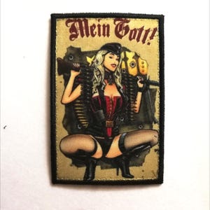 Peut inclure: Écusson brodé représentant une femme de style pin-up en tenue noire, tenant une mitrailleuse. L'écusson porte les mots "Mein Gott!" dans une police décorative, sur fond beige avec une bordure noire.