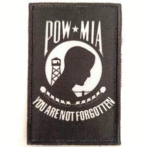 Peut inclure: Écusson rectangulaire noir avec texte blanc et silhouette de profil. Le texte indique "POW*MIA" au-dessus d'un motif circulaire avec une tour de guet et les mots "YOU ARE NOT FORGOTTEN".