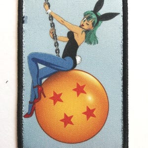 May include: Embroidered patch showing a cartoon woman with green hair and bunny ears, sitting on an orange ball with four red stars. She wears a black top, blue trousers, and red heels, holding a chain.