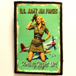 Peut inclure: Un écusson de style vintage représentant une femme blonde en uniforme militaire posant sur un avion. L'écusson porte les inscriptions "U.S. ARMY AIR FORCES" et "COMING Right UP!" sur fond vert, encadré d'une bordure noire.