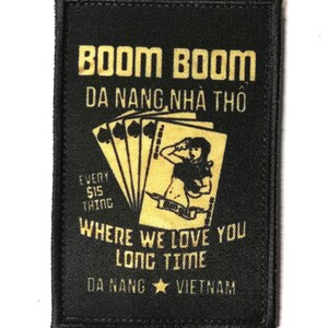 May include: Black rectangular patch with gold text and imagery. The patch reads "BOOM BOOM DA NANG, NHÀ THÔ" and "WHERE WE LOVE YOU LONG TIME DA NANG VIETNAM." A stylized image of playing cards and a woman is also present.