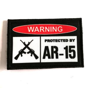 Könnte beinhalten: Schwarz-weißer Aufnäher mit dem Text "WARNING" in einem roten Oval und "PROTECTED BY AR-15". Mit gekreuzten Gewehren. Der Aufnäher hat einen schwarzen Rand.