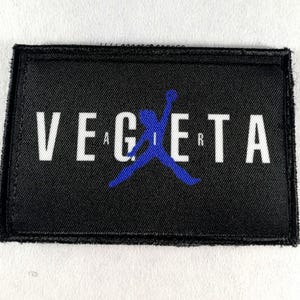 May include: Black rectangular patch with the word "VEGETA" in white capital letters. A blue silhouette of a person jumping is in the center. The patch has a black border and is on a white background.