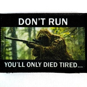 Peut inclure: Écusson rectangulaire noir avec le texte "DON'T RUN YOU'LL ONLY DIED TIRED...". L'écusson présente une image d'un tireur d'élite camouflé visant avec un fusil dans un cadre forestier. Le texte est en blanc.