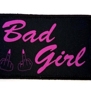 Op de afbeelding: Zwart rechthoekig embleem met de woorden "Bad Girl" in roze script. Twee roze omrande middelvingers staan onder de tekst. Het embleem heeft een zwarte rand.
