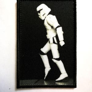 Puede incluir: Parche en blanco y negro con un Stormtrooper de Star Wars. El parche tiene un fondo negro y un borde cosido. El Stormtrooper se representa en una pose de caminar.
