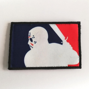 Puede incluir: Un parche rectangular con una silueta blanca de máscara de hockey y una figura sosteniendo un machete, sobre un fondo azul marino y rojo. El parche tiene un borde negro.