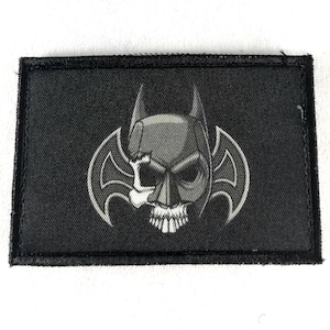 Peut inclure: Écusson rectangulaire noir avec un motif de crâne gris et des ailes de chauve-souris de style Batman. Le crâne est partiellement visible. L'écusson a une bordure noire.