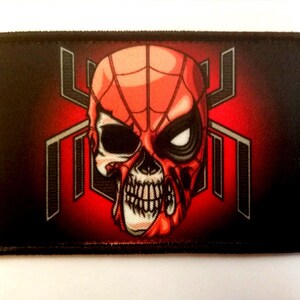 Könnte beinhalten: Ein rechteckiger Aufnäher mit einem rot-schwarzen Design. Das Bild zeigt einen Schädel mit einer Spider-Man-Maske, vor einem roten Hintergrund mit einem stilisierten Spinnennetz-Design. Der Aufnäher hat einen schwarzen Rand.