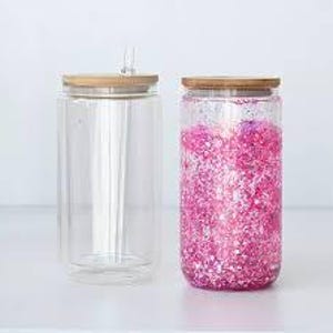 May include: Two clear glass tumblers with bamboo lids. One tumbler is empty with a straw, the other is filled with pink glitter. The tumblers are cylindrical and sit on a white surface.