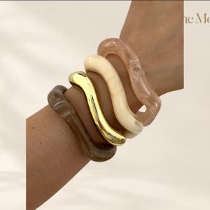 Op de afbeelding: Een set van vier golvende armbanden die samen worden gedragen. De set bevat een doorschijnende bruine harsarmband, een gepolijste goudkleurige metalen armband, een ondoorzichtige crèmekleurige harsarmband en een doorschijnende lichtroze harsarmband. Tekst "the Moft".