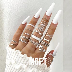 Könnte beinhalten: Eine Sammlung silberner Ringe in verschiedenen Designs, darunter Bänder, Ketten und Blumenmotive, wird auf Fingern mit langen, weißen Nägeln präsentiert. Die Ringe werden an mehreren Fingern getragen und erzeugen einen mehrschichtigen Look. Das Wort "MOFT" ist sichtbar.