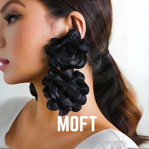 Può includere: Orecchini a fiore neri oversize. Ogni orecchino presenta due grandi fiori di tessuto a strati. Gli orecchini sono un pezzo audace, perfetto per aggiungere drammaticità a qualsiasi outfit. La parola "MOFT" è stampata in bianco.
