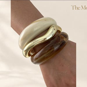 Puede incluir: Un conjunto de tres brazaletes apilados que incluye una pulsera de resina color crema lisa, una pulsera de metal dorado pulido con silueta ondulada y una pulsera de resina marrón translúcida. El texto The Moft es visible en la esquina.