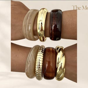 Può includere: Una collezione di sette bracciali rigidi indossati su due polsi. Il set include resina beige liscia, metallo dorato lucido, resina traslucida marrone scuro, un bracciale in metallo dorato a coste e uno intrecciato. Il testo riporta The Moft.