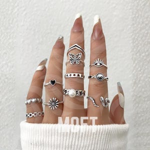 May include: A selection of silver rings featuring diverse designs such as hearts, butterflies, and a daisy. The rings are presented on fingers, highlighting a range of styles and sizes. The word "MOFT" is visible in the image.