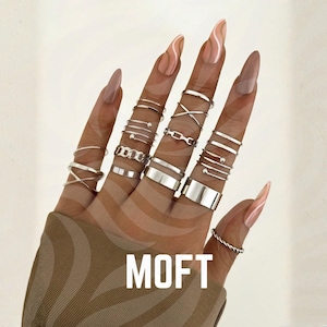 Könnte beinhalten: Eine Sammlung silberner Ringe in verschiedenen Designs, darunter Bänder, Kettenglieder und Kreuzmuster. Die Ringe werden auf einer Hand mit langen, gepflegten Nägeln präsentiert. Das Wort "MOFT" ist auf dem Bild aufgedruckt.