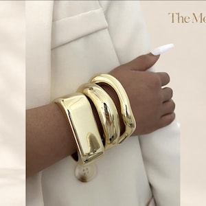 Può includere: Un set di tre bracciali rigidi in metallo color oro con una finitura lucida e riflettente. I bracciali presentano larghezze variabili e contorni lisci e arrotondati. Il testo "The Moft" è visibile nell'angolo in alto a destra.