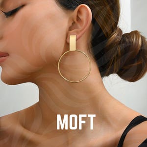 May include: Gold-colored hoop earrings with a rectangular top piece. The earrings are circular and hang from the ear. The word "MOFT" is printed in white block letters at the bottom of the image.