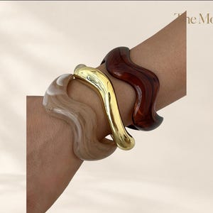 Può includere: Tre bracciali rigidi ondulati indossati insieme. Un bracciale è in metallo color oro lucido, uno è in resina marrone scuro traslucida e uno è in resina beige marmorizzata. Ogni bracciale presenta una forma curva e ondulata con finitura liscia.