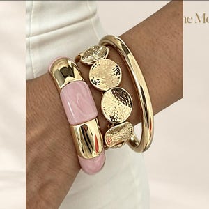 Puede incluir: Un conjunto de tres pulseras que incluye un brazalete liso rosa y dorado, una pulsera dorada con discos circulares martillados y un brazalete dorado pulido. El texto "the Moft" es visible en la esquina superior derecha.