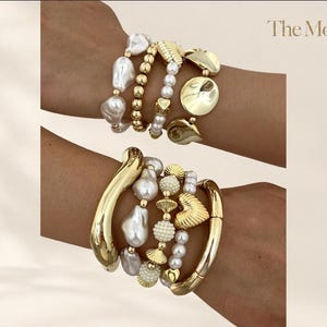 Può includere: Due set di bracciali sovrapposti con perle barocche bianche, perline metalliche dorate e dischi dorati lisci. Un set include un grande bracciale rigido dorato curvo e un ciondolo a forma di cuore testurizzato. Il testo The Moft è visibile.