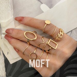 Pode incluir: Uma coleção de anéis dourados em vários designs, incluindo um círculo, um oval, um quadrado e um padrão de folhas. Os anéis são exibidos em uma mão com unhas cuidadas. A palavra "MOFT" é visível.