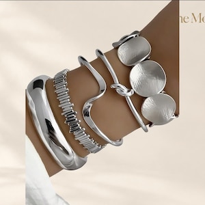 Puede incluir: Un conjunto de cuatro pulseras plateadas que incluye un brazalete ancho y liso, una cadena de eslabones de barra texturizados, una banda de alambre ondulado pulido y una banda de alambre anudado con tres discos circulares. El texto "the Moft" es visible arriba a la derecha.