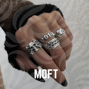 Peut inclure: Quatre bagues en argent sont présentées sur une main, avec des motifs de crâne et d'os croisés, de crâne et de corde. Les bagues sont finement détaillées et portées sur différents doigts. Le mot "MOFT" est visible en blanc en bas de l'image.